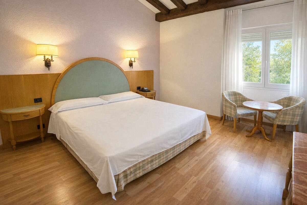 A hotel room with a bed, bedside tables, a chair, a small table, and a window with curtains.