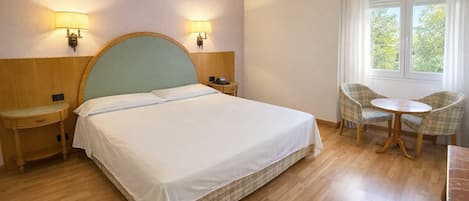 A hotel room with a bed, bedside tables, a chair, a small table, and a window with curtains.