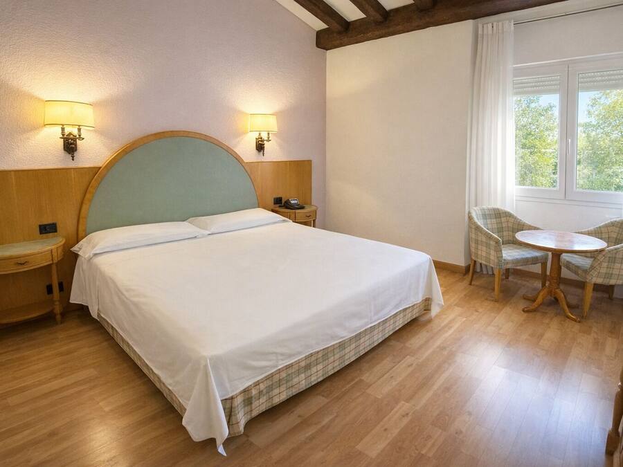 Habitación de hotel con cama, mesitas de noche, una silla, una mesita pequeña y una ventana con cortinas.