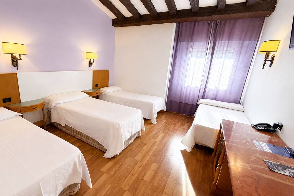 A hotel room with three single beds, wooden headboards, a wooden desk, and two wall-mounted lamps.