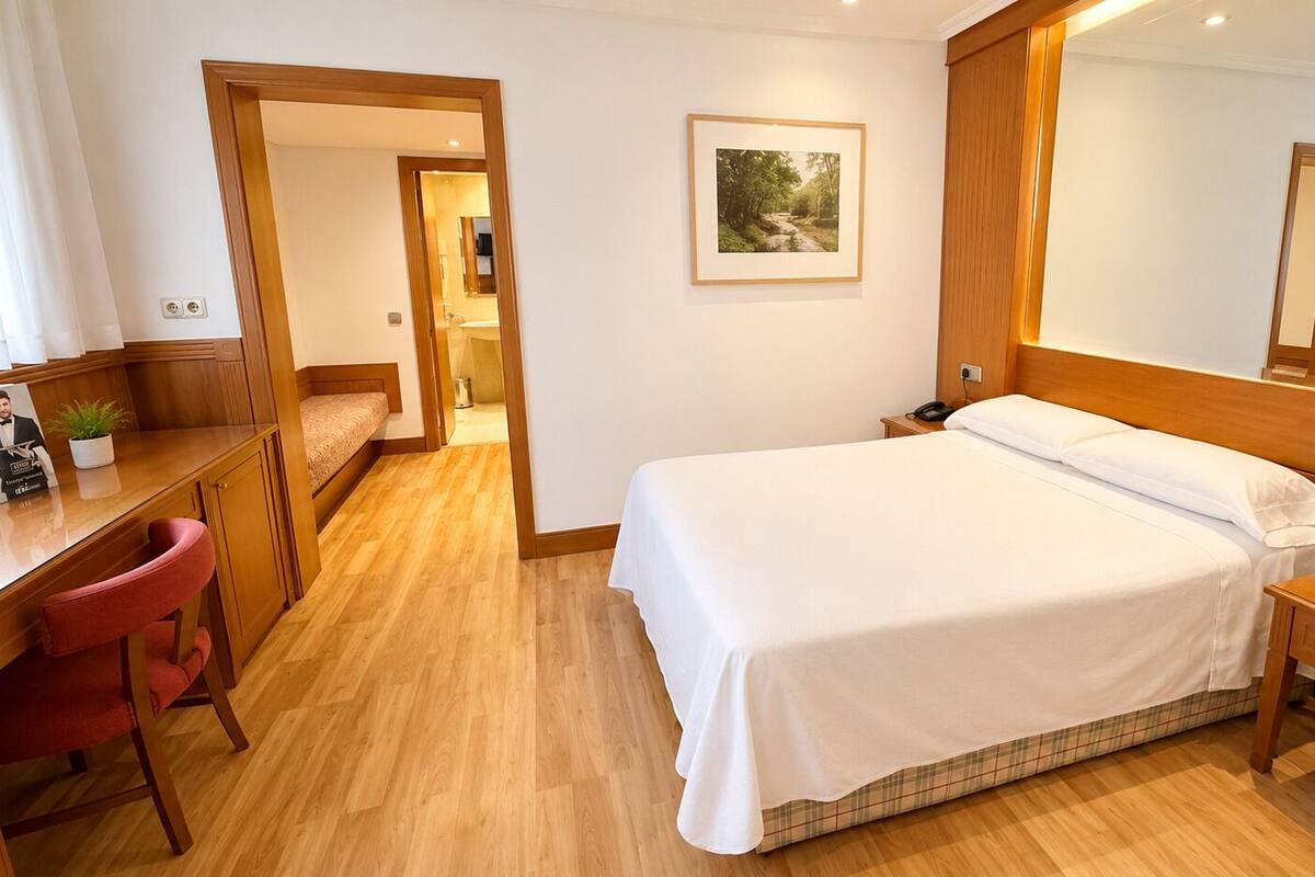A hotel room with a bed, a desk, a chair, a bedside table, a mirror, and a framed picture on the wall.