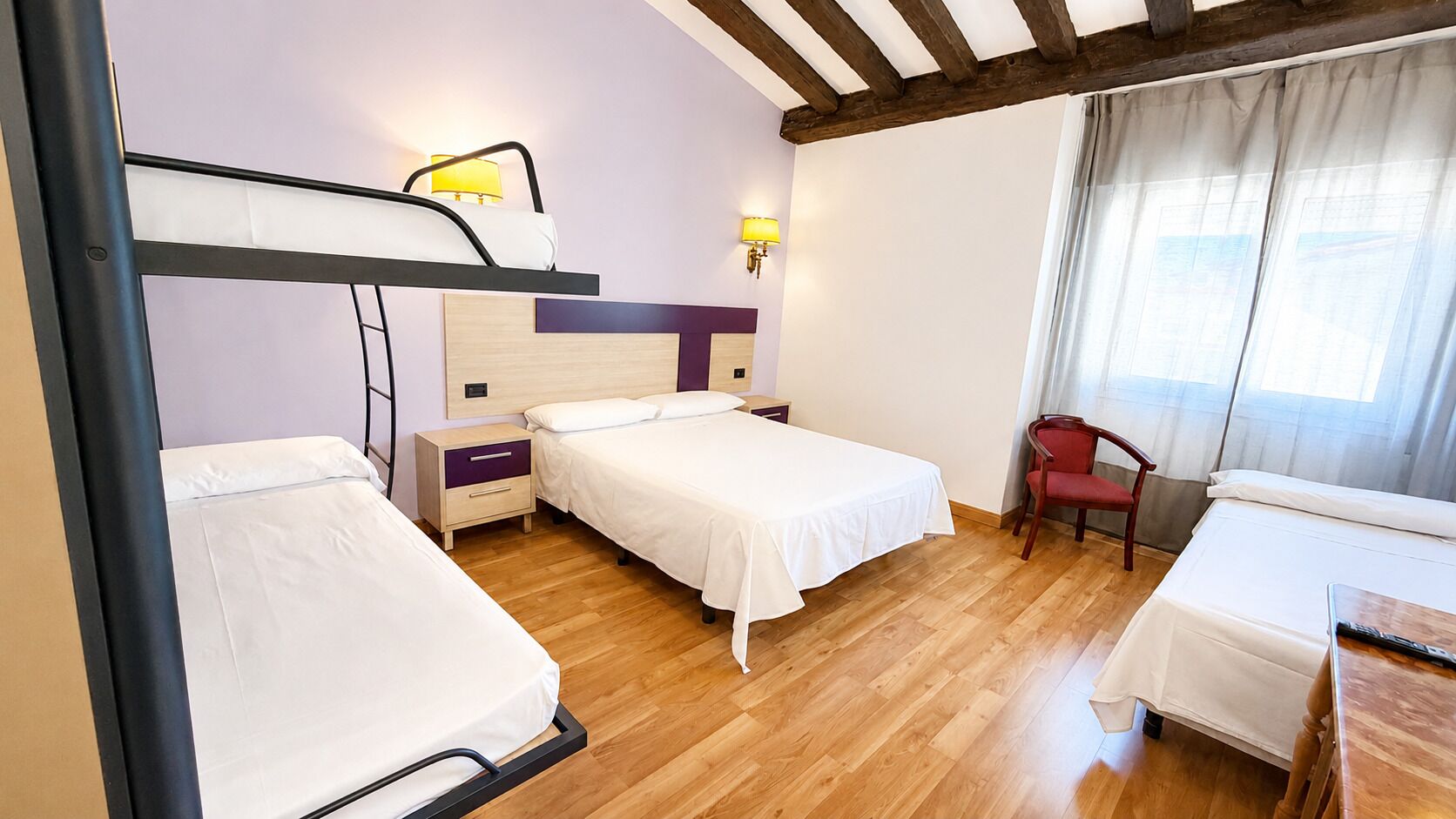 A room with two beds, a wooden floor, a desk with a chair, and a window with curtains.