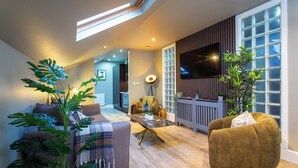 A modern living room with a sofa, armchair, and coffee table, a large window with blinds, and a flat-screen TV mounted on the wall. - Luxury apartment & terrace view over river & easy access to City Centre & Castle (Inverness)