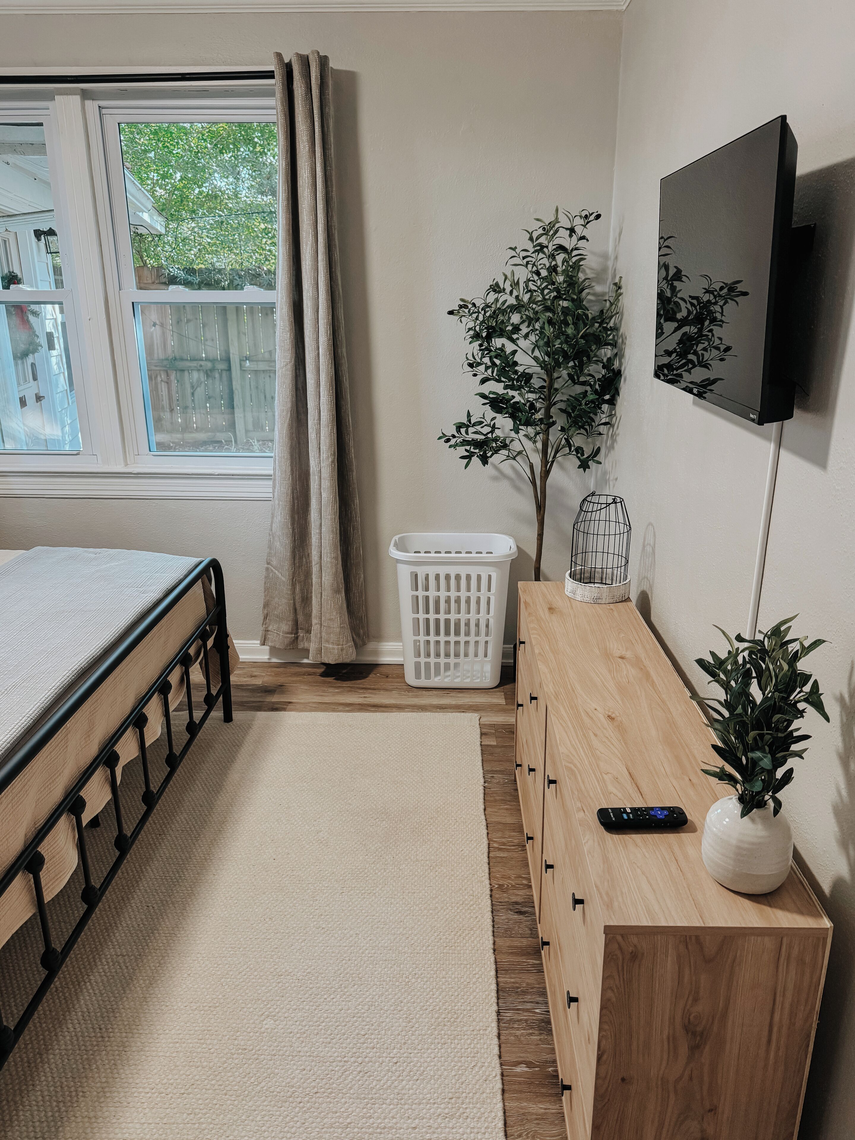 A bedroom with a bed, a dresser, a TV, a plant, and a laundry basket.
