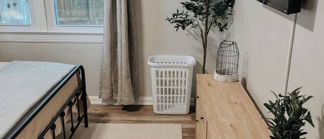 Un dormitorio con cama, cómoda, televisor, planta y cesto de ropa.