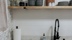 Una cocina con encimera de mármol, pileta de acero inoxidable, grifo negro, dispensador de toallas de papel, una botella de jabón para manos y una estantería con vajilla y una planta.