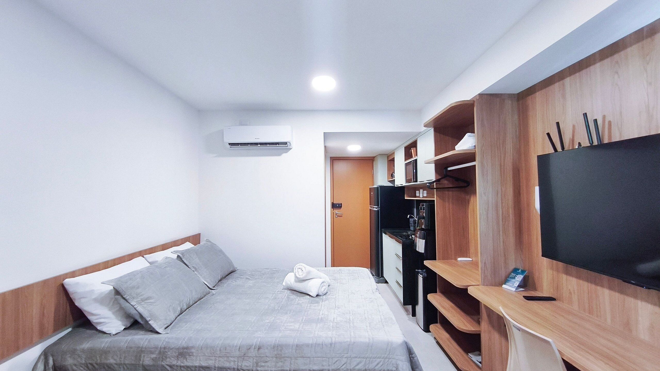 A compact hotel room with a bed, a TV, a wooden wardrobe, and a kitchenette.