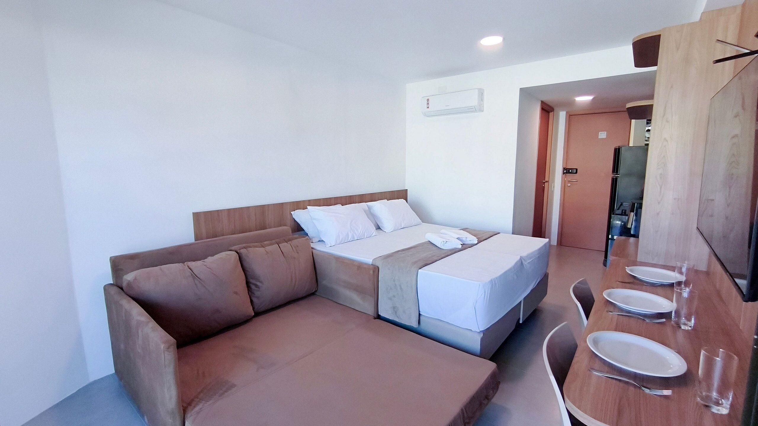Quarto de hotel moderno com sofá-cama, mesa de jantar com pratos e talheres, e porta visível para outro cômodo.