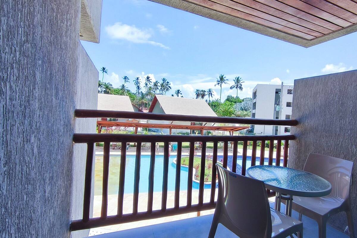 A balcony with a view of a pool and palm trees, a table, and chairs.