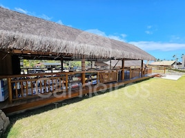 Un restaurante frente a la playa con techo de paja, terraza de madera y mesas al aire libre.