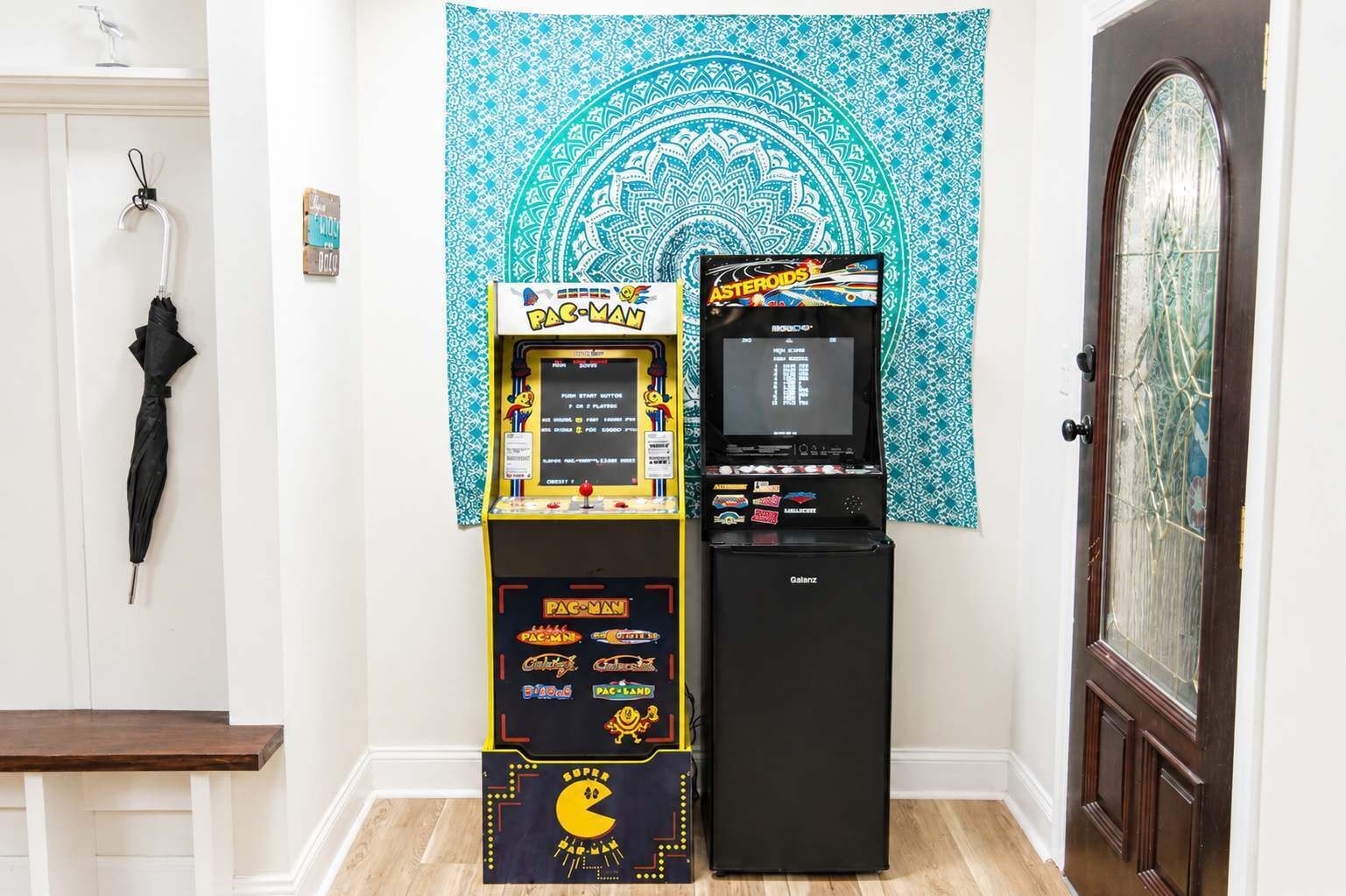 Una máquina recreativa de Pac-Man y un refrigerador Galanz en una habitación con una puerta, un perchero y un tapiz.