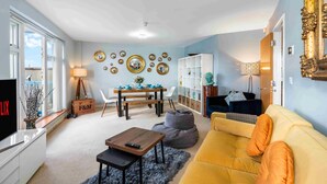 A living room with a sofa, a coffee table, a TV, a dining table with chairs, and a bookshelf. - River Heights With Views Parking |By My Getaways (Brighton)