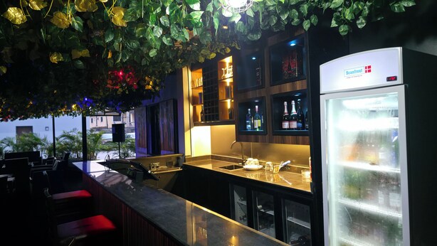 Een bar met een toog, planken vol flessen en een koelkast.