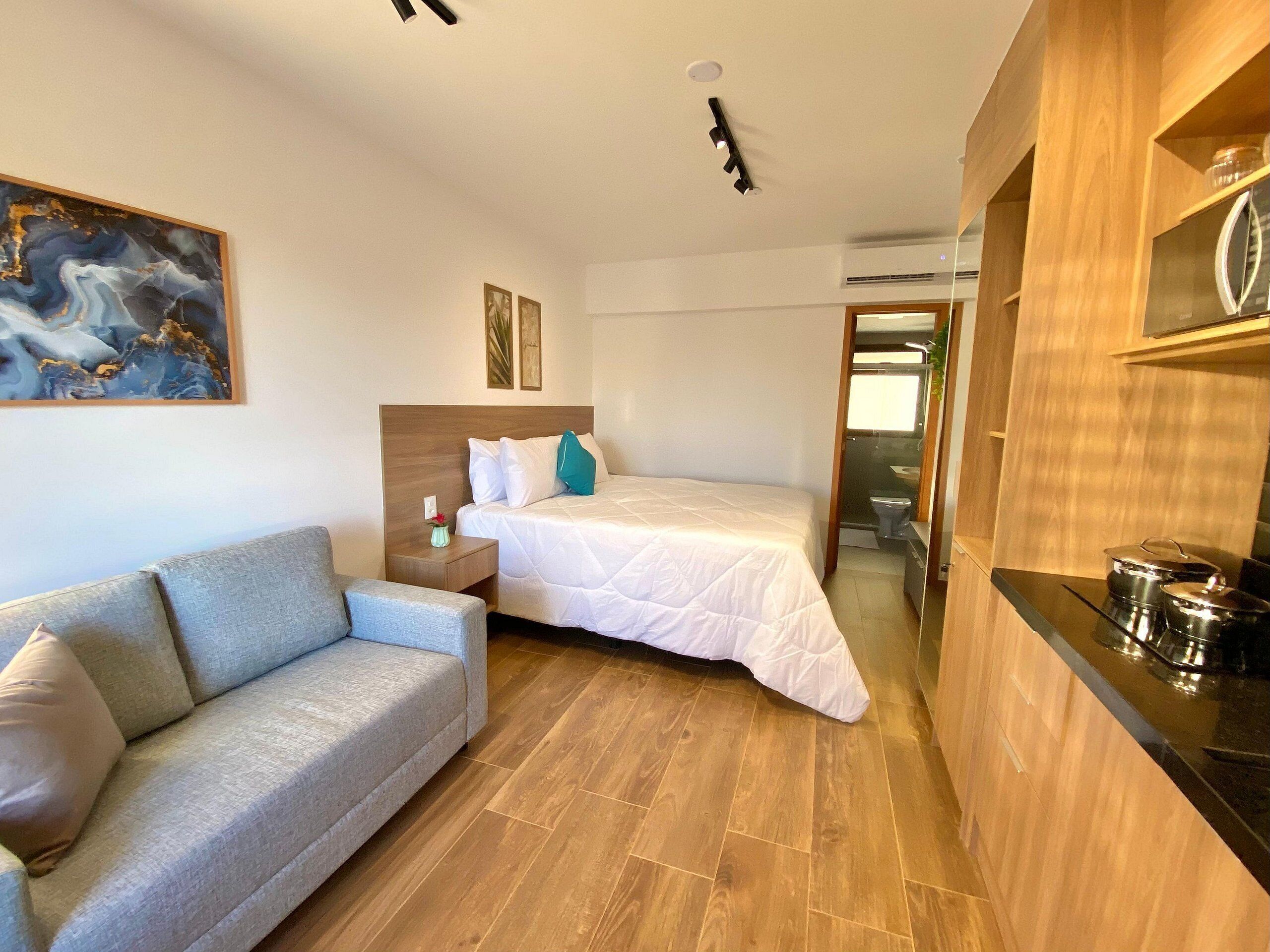 A compact hotel room with a bed, a sofa, a small table, a painting, and a kitchenette.