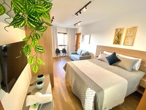 A hotel room with a bed, a TV, a desk, two chairs, and a potted plant.