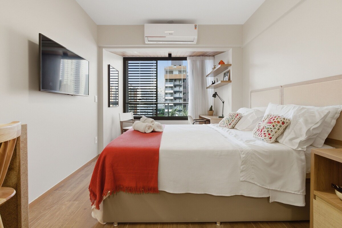 Ein Schlafzimmer mit Bett, Fernseher, Fenster mit Jalousien und Blick auf Gebäude.