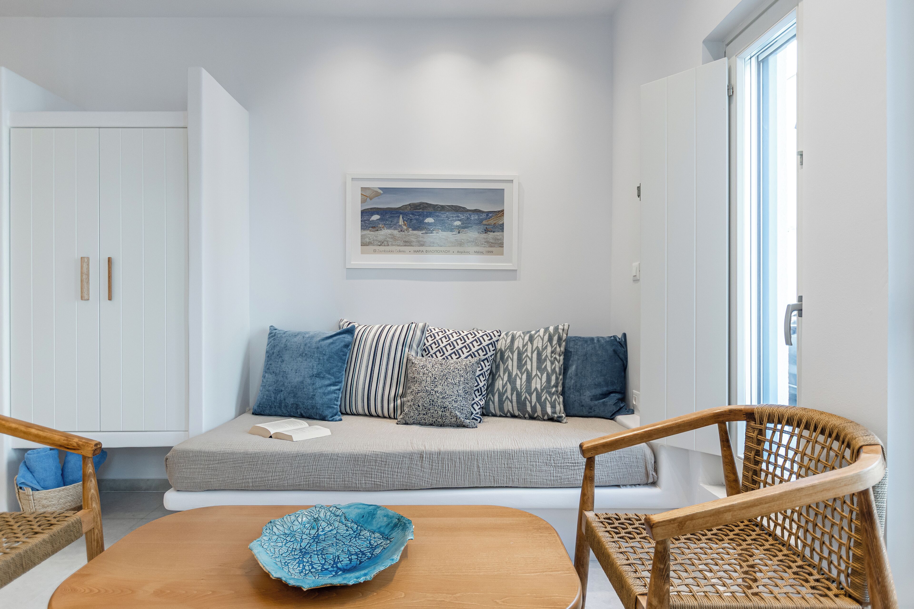 A modern living room with a sofa, two wicker chairs, a wooden table with a blue bowl, and a framed painting on the wall.