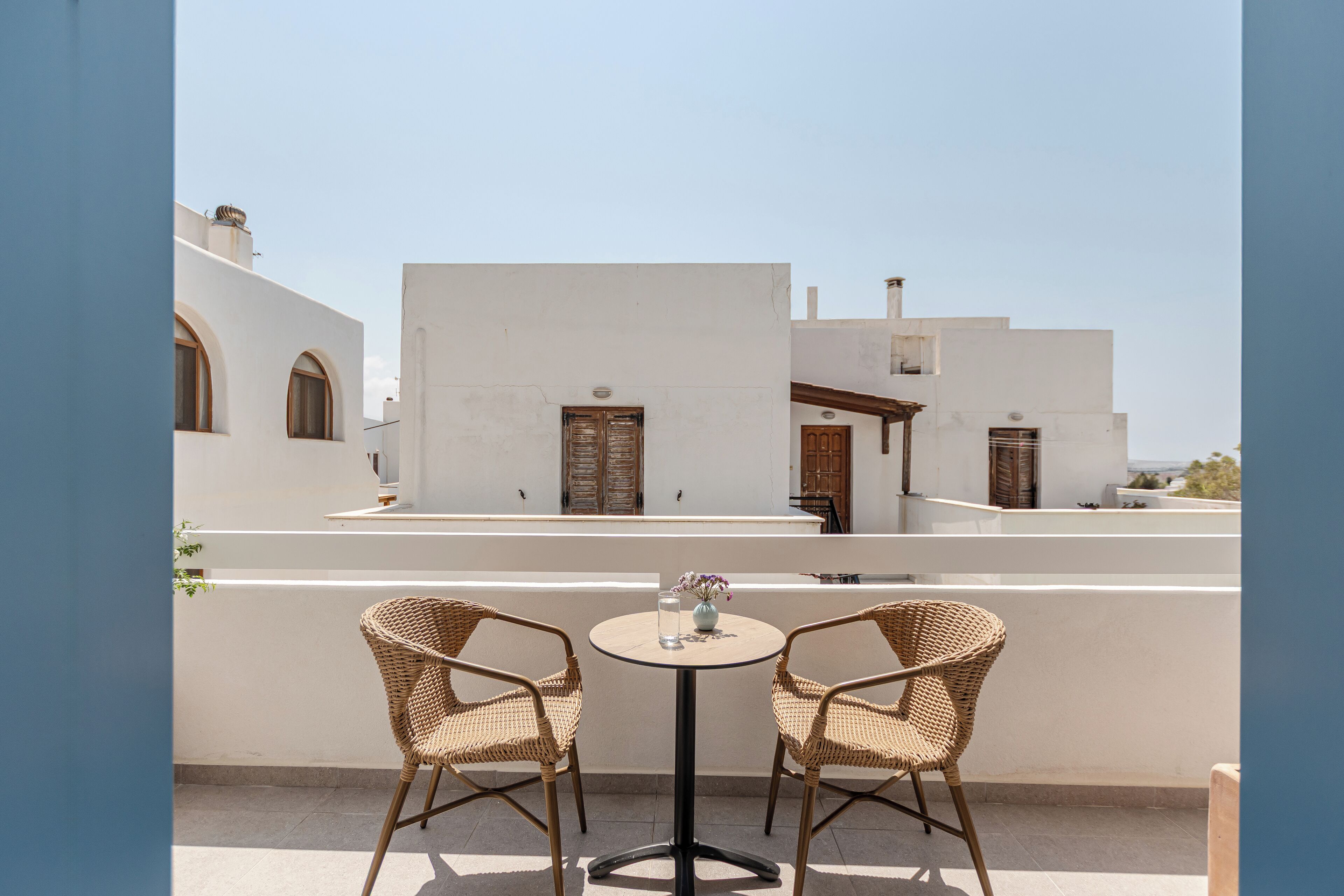 Eine Terrasse mit zwei Korbstühlen und einem kleinen runden Tisch, mit Blick auf ein weißes Gebäude mit Rundbogenfenstern und Holztüren.