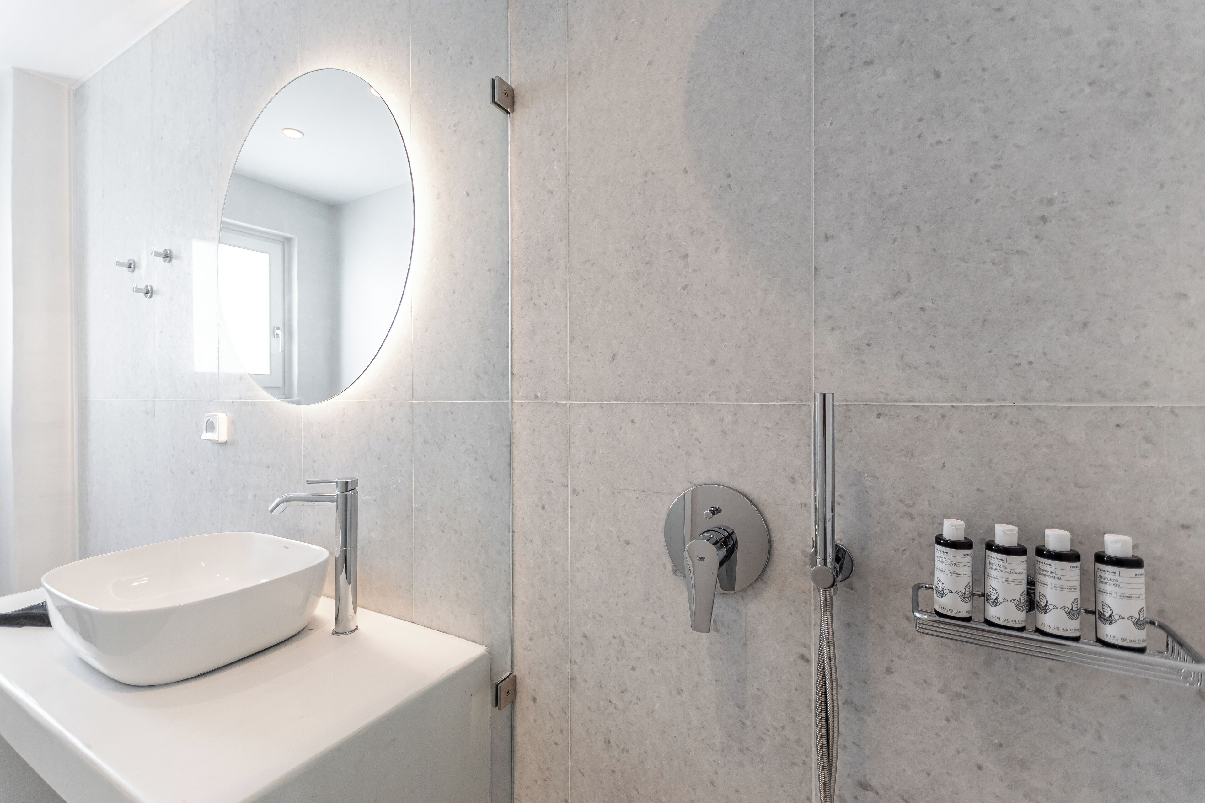 A modern bathroom with a white sink, a round mirror, a handheld showerhead, and a shelf with three bottles.