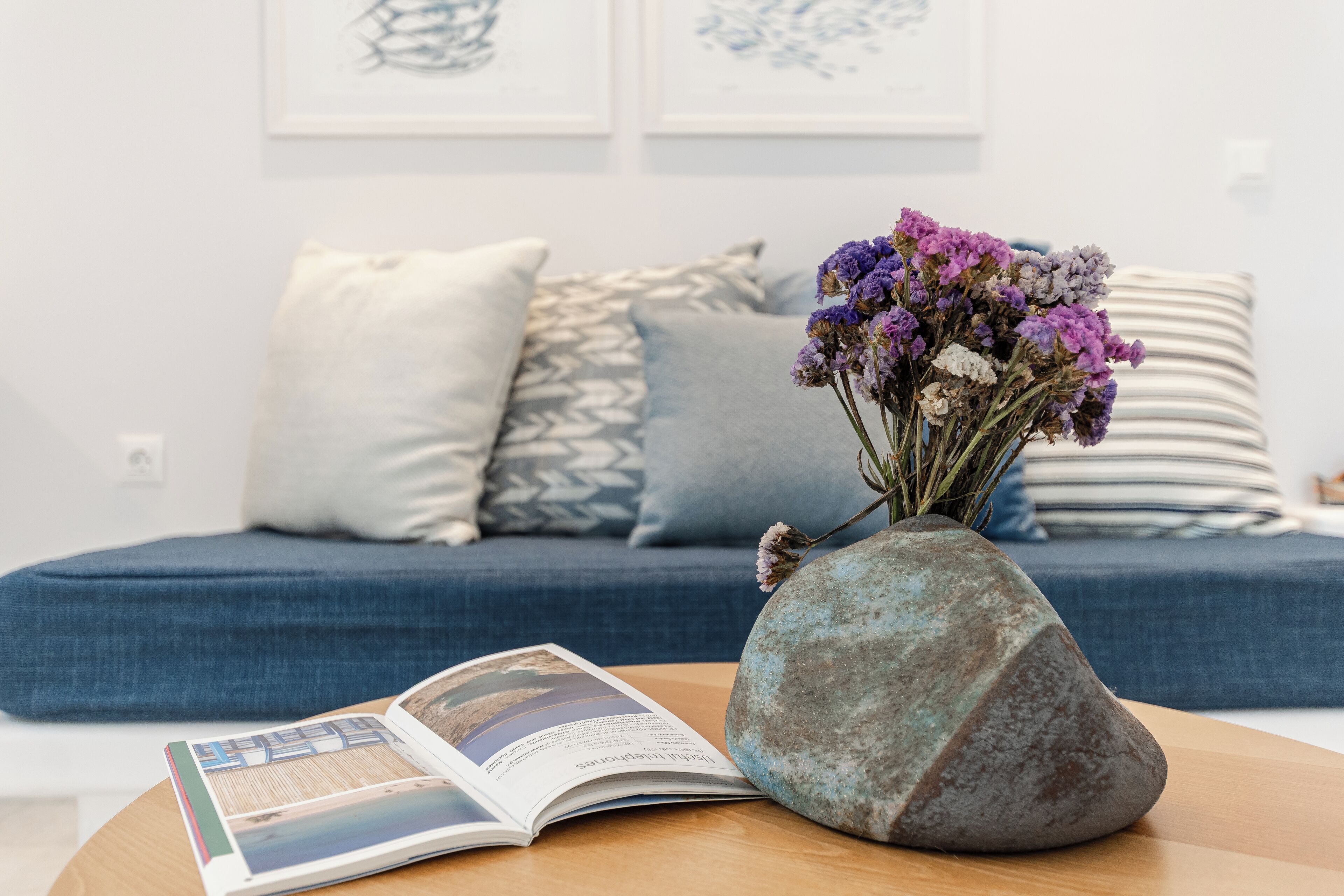 A room with a blue sofa, a stone vase with flowers, and an open magazine on a wooden table.