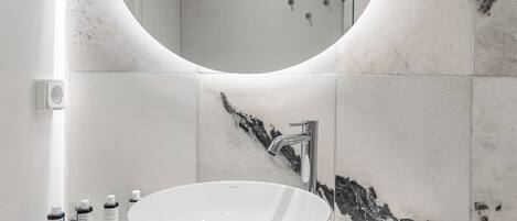 Un baño moderno con un espejo redondo, lavamanos blanco y azulejos de mármol.