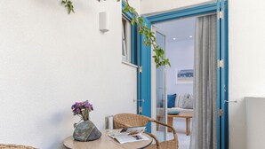 Ein kleiner runder Tisch mit einer Vase voller lila Blumen, ein Korbstuhl und eine Zeitschrift. Eine blaue Tür führt zu einem Zimmer mit einem Bett und einem Fenster.