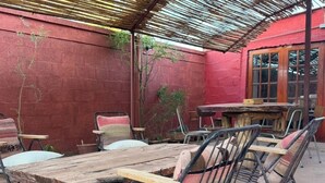 Une terrasse extérieure rustique, avec une table en bois et des chaises en métal, entourée de murs rouges et surmontée d’une pergola en bambou.