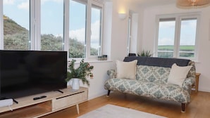 Una sala de estar con un televisor de pantalla plana grande sobre un soporte de madera, un sofá estampado, una alfombra blanca y vistas al exterior a través de amplios ventanales.