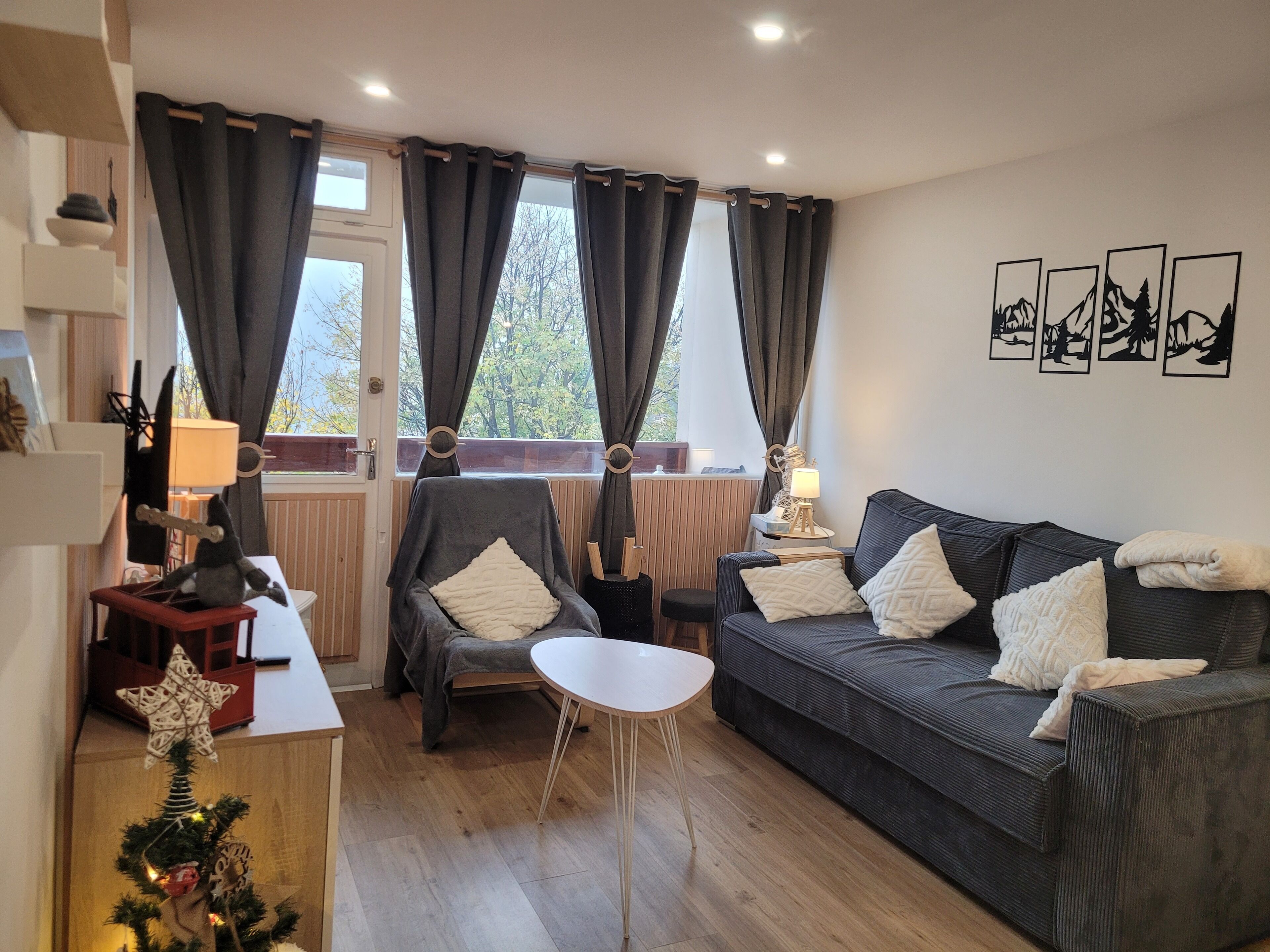 Ein gemütliches Wohnzimmer mit einer Couch, einem Sessel und einem kleinen runden Tisch. Vorhänge, ein Fenster mit Blick auf Bäume und eine Wand mit gerahmten Bildern sind ebenfalls vorhanden.
