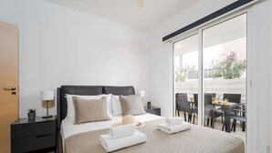 A modern hotel room with a large bed, a bedside table with a lamp, a small desk with a chair, and a balcony with a dining table and chairs. - Stylish & Bright Gem ~ Walk to Beach, Patio, Pools (Paralimni)