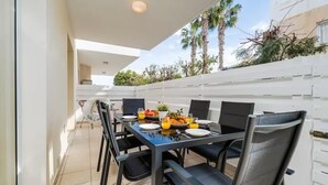 A dining area with a table set for four, outdoor chairs, and a view of palm trees and a white wall. - Stylish & Bright Gem ~ Walk to Beach, Patio, Pools (Paralimni)