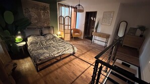 A bedroom with a bed, a nightstand, a chair, a small table, a mirror, and a staircase. - The little refuge (Quimperlé)