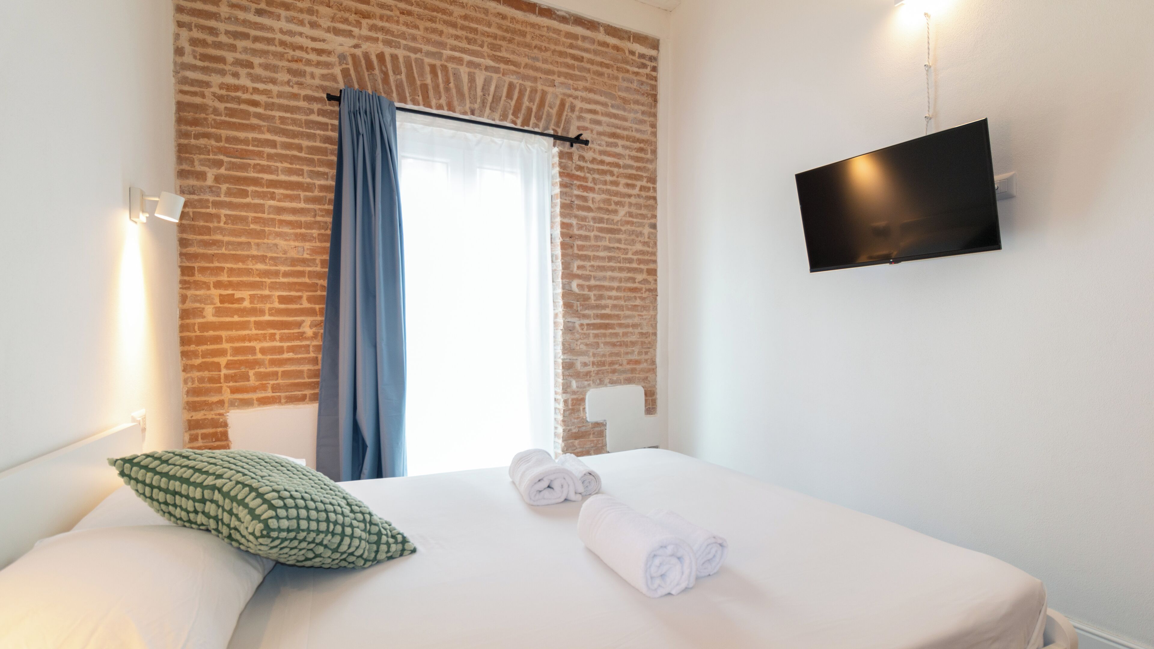 Quarto de hotel com parede de tijolos, cama com roupa de cama branca, travesseiro verde e cortina azul, além de uma TV de tela plana fixada na parede.