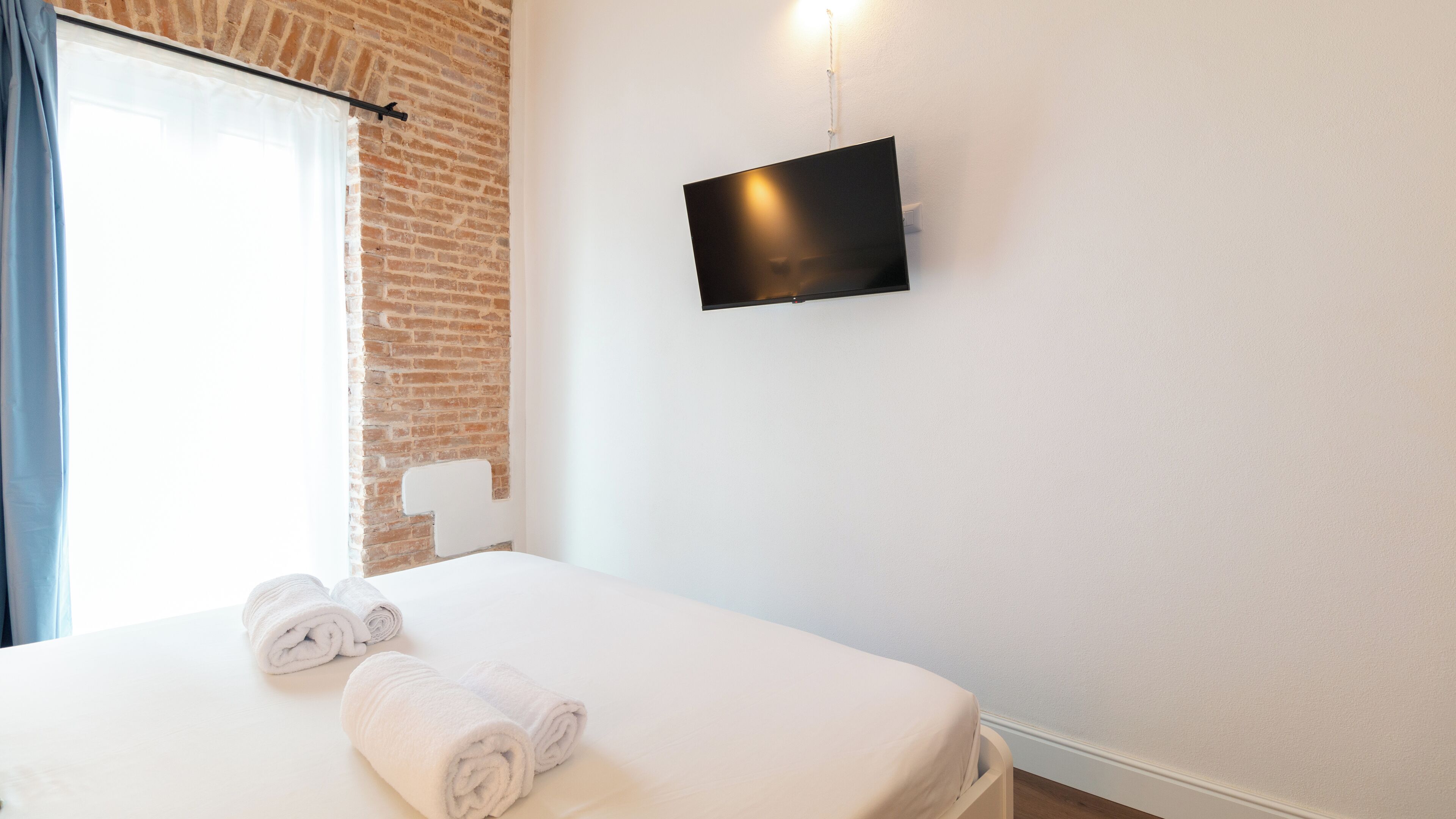 A hotel room with a bed, a flat-screen TV mounted on the wall, and a window with curtains.