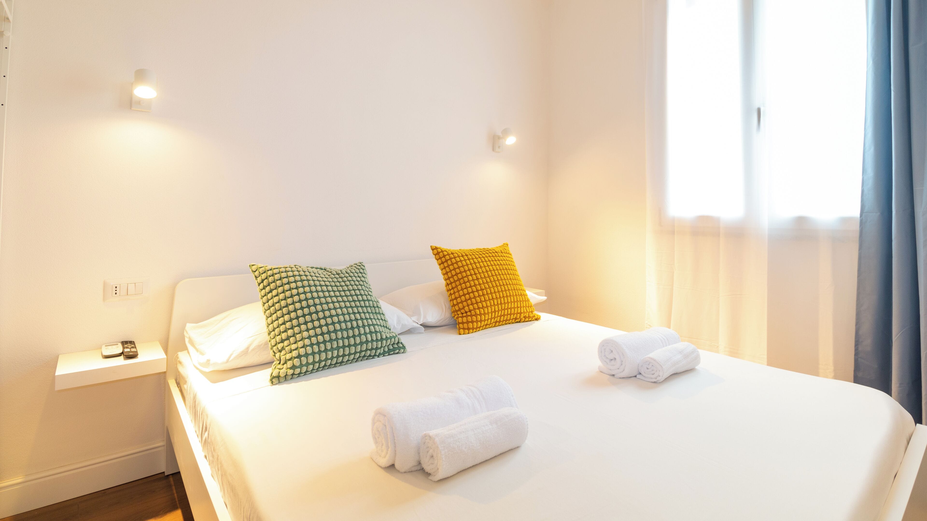 A hotel room with a bed, two pillows, a nightstand, a remote control, and two towels.