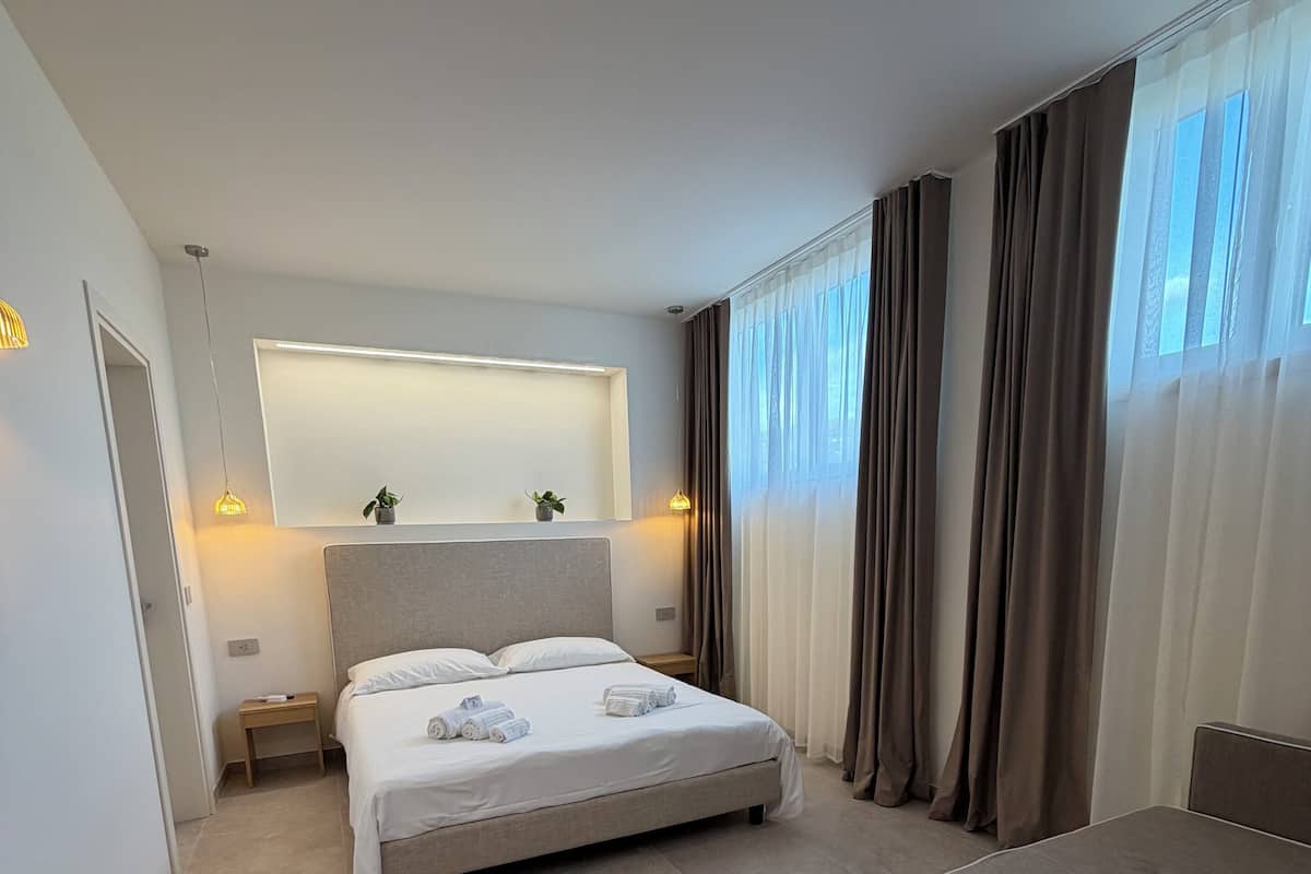 A modern hotel room with a large bed, a bedside table, a small table, and a large window with curtains.