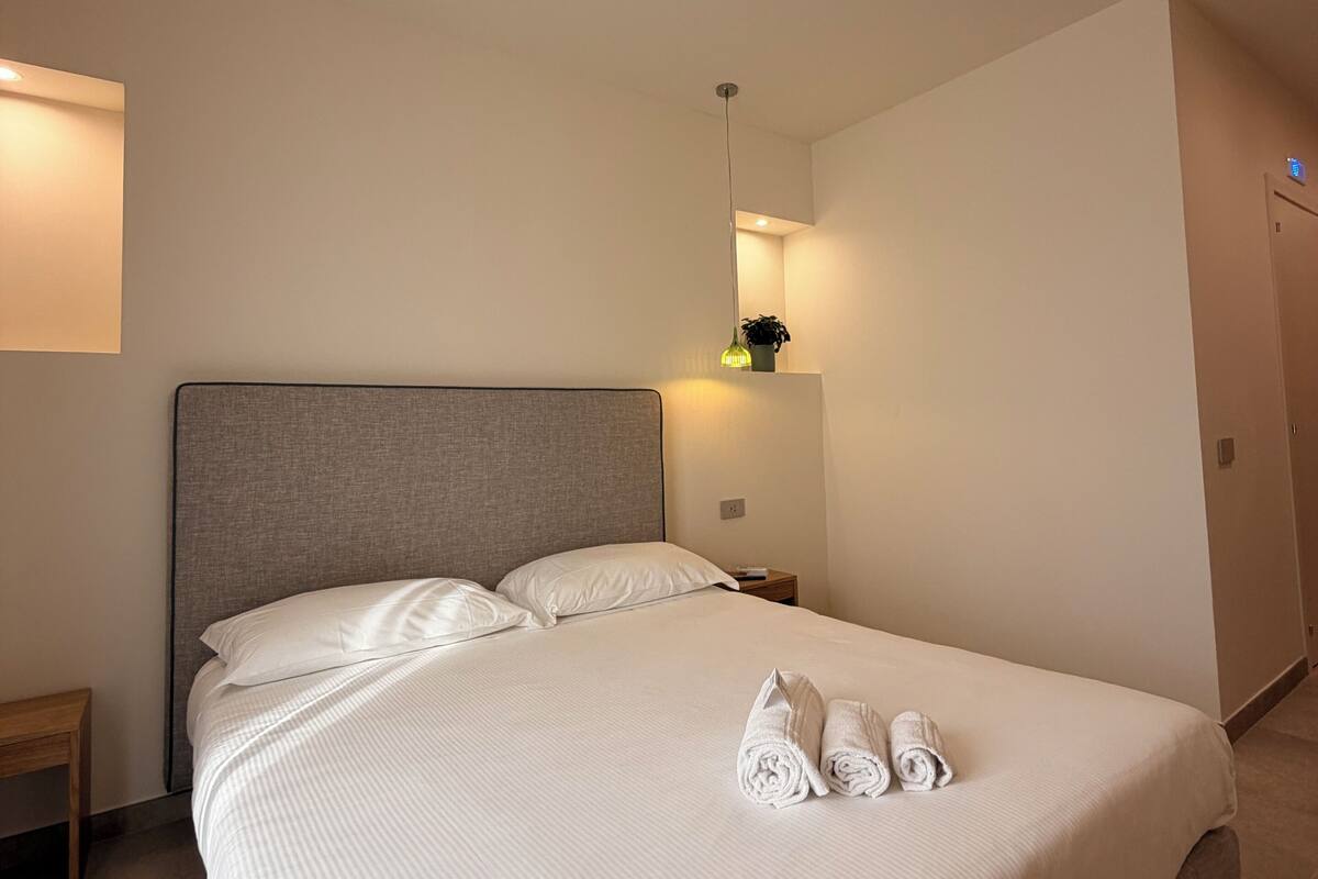 A neatly made bed with white linens and rolled towels, a bedside table with a lamp, and a wall-mounted shelf with a plant.