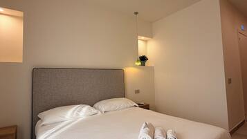 A neatly made bed with white linens and rolled towels, a bedside table with a lamp, and a wall-mounted shelf with a plant.