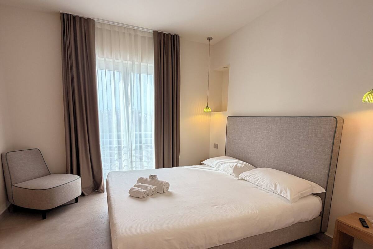 A neatly arranged hotel room with a large bed, a chair, a bedside table, and a window with curtains.