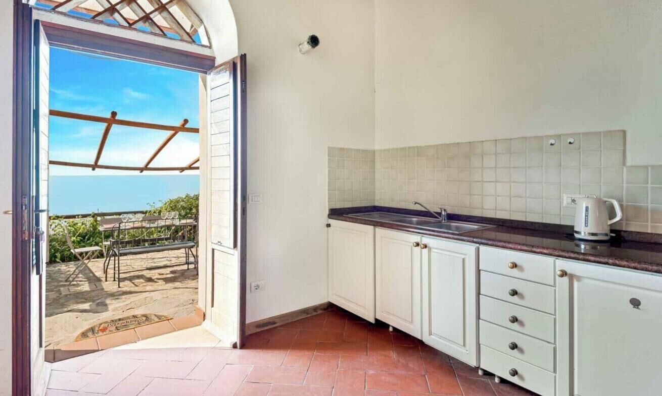 Et køkken med hvide skabe, et flisebeklædt bagpanel og udsigt gennem en åben dør til et udendørs område med et pergola.