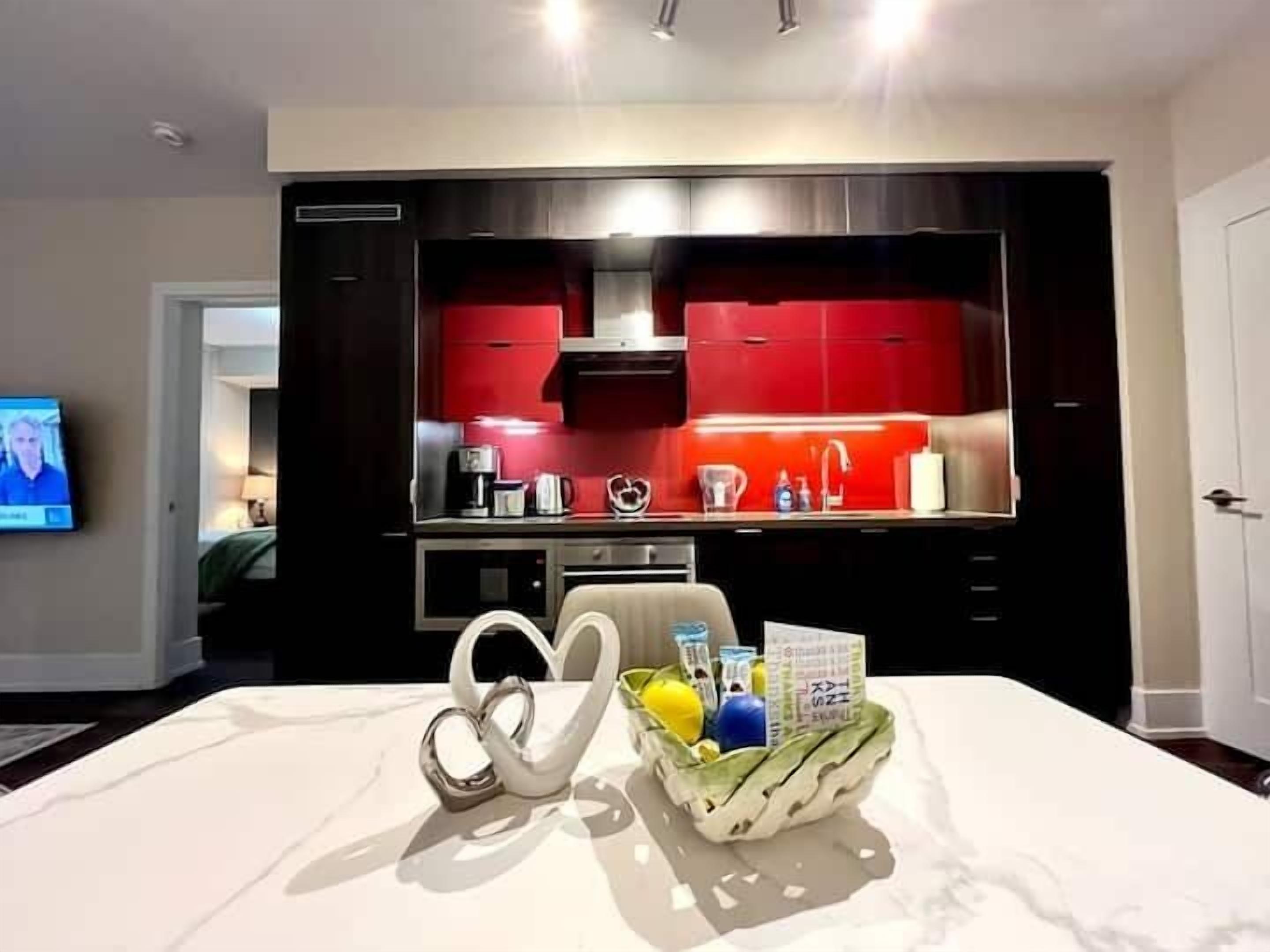 A modern kitchen with red cabinets, a microwave, and a coffee maker. There is a dining table with a white tablecloth and a fruit basket.