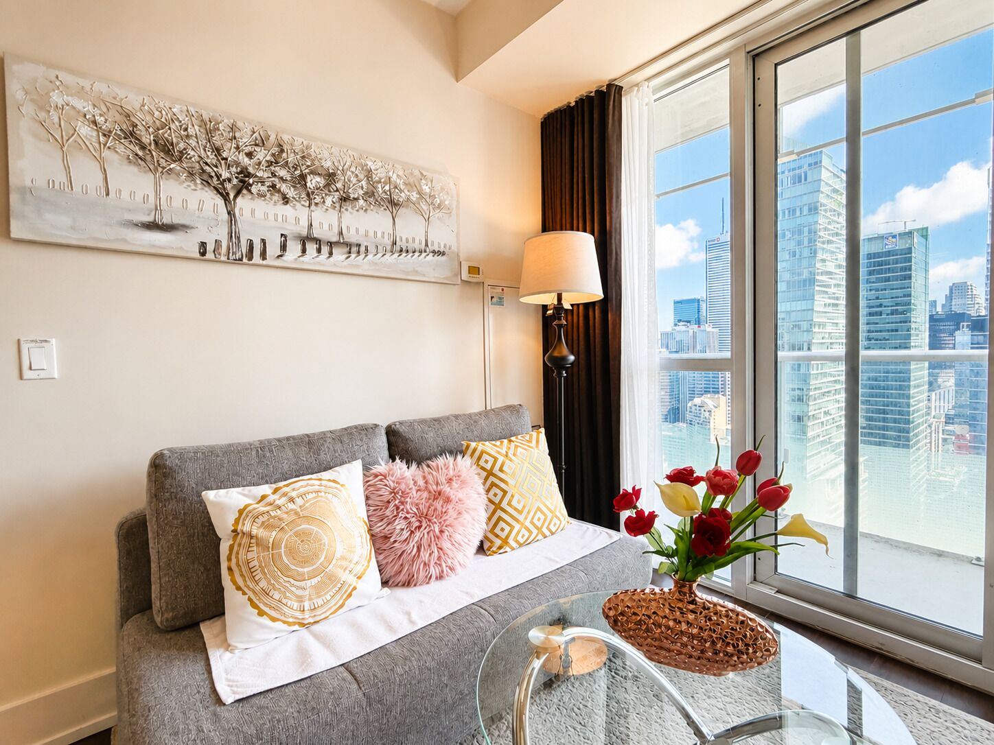 Een moderne woonkamer met een grijze bank, decoratieve kussens, uitzicht op de stad en een wandlamp.