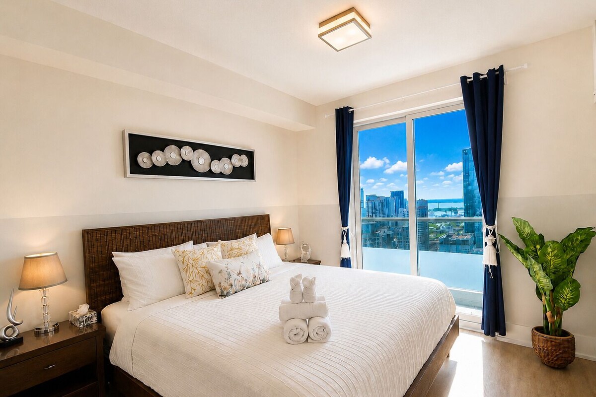 Une chambre d’hôtel avec un grand lit, une table de chevet, une lampe, un tableau décoratif au mur, une fenêtre avec des rideaux et une vue sur la ville.