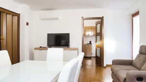 A modern living room with a white dining table, a beige sofa, and a flat-screen TV on a wooden stand.