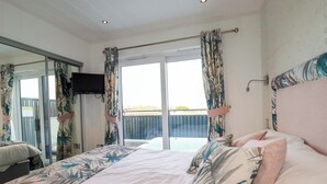 A bedroom with a large bed, a TV mounted on the wall, and a sliding glass door leading to a balcony. - South Crofty - The Residence (Redruth)