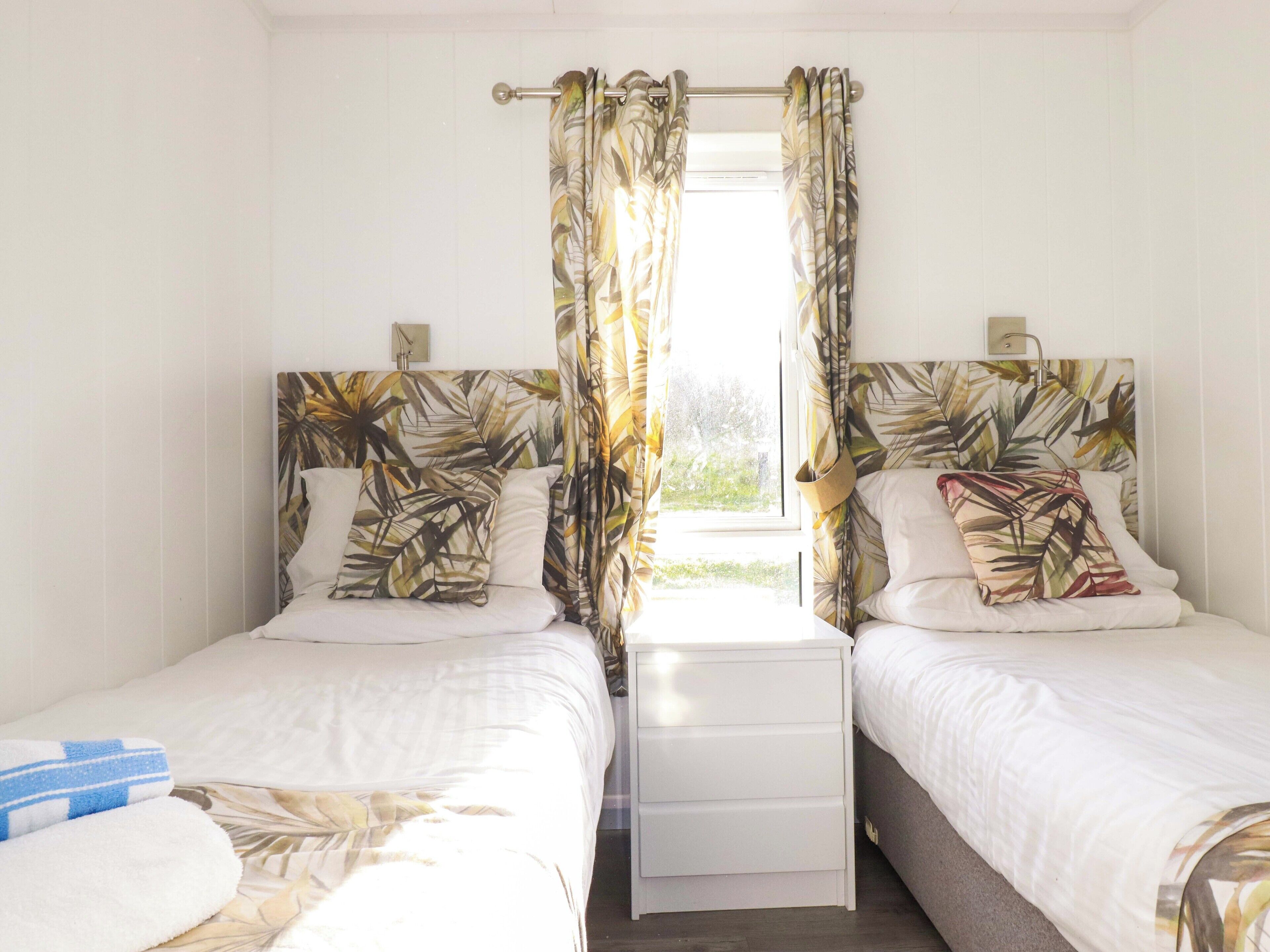 A double room with two beds, each with a headboard, a bedside table, and a window with curtains.