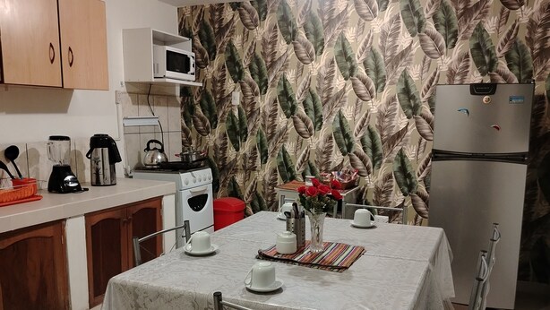 Una pequeña cocina con armarios de madera, un microondas, un refrigerador y una mesa de comedor dispuesta con tazas y un jarrón de flores.