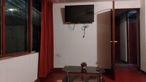 Una habitación pequeña con un televisor en la pared, una mesa con un florero y un control remoto, y una puerta que da a otra habitación.