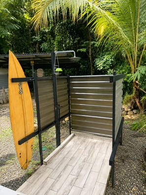 Een houten terras met een metalen hek, een surfplank tegen het hek leunend en een douchekop erboven.