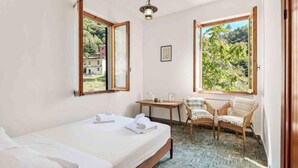 Ein Schlafzimmer mit einem Bett, zwei Stühlen, einem kleinen Tisch und einem Fenster mit Blick auf Grünflächen.